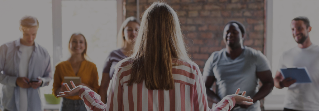 A woman with her back to the camera speaks with her arms open to a diverse group of smiling people in a loft-style room.