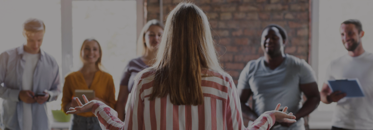 A woman with her back to the camera speaks with her arms open to a diverse group of smiling people in a loft-style room.