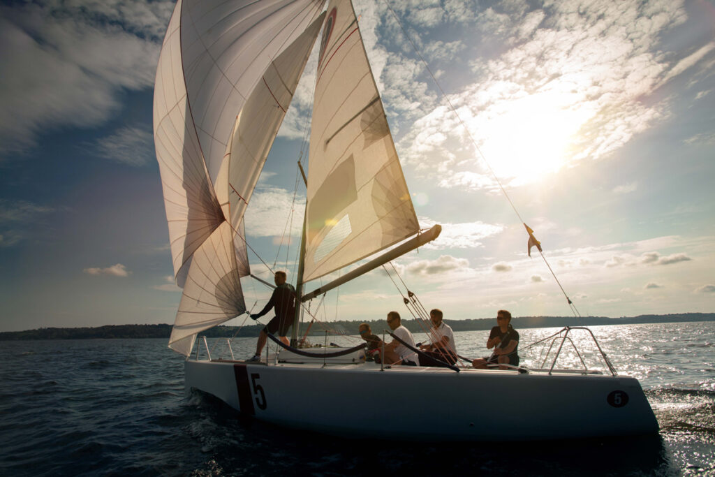 A crew sailing a sailboat with full sails across sunny open water.