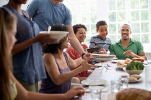 **Alt Text:** A diverse group of smiling people sharing and passing dishes around a dining table filled with food.