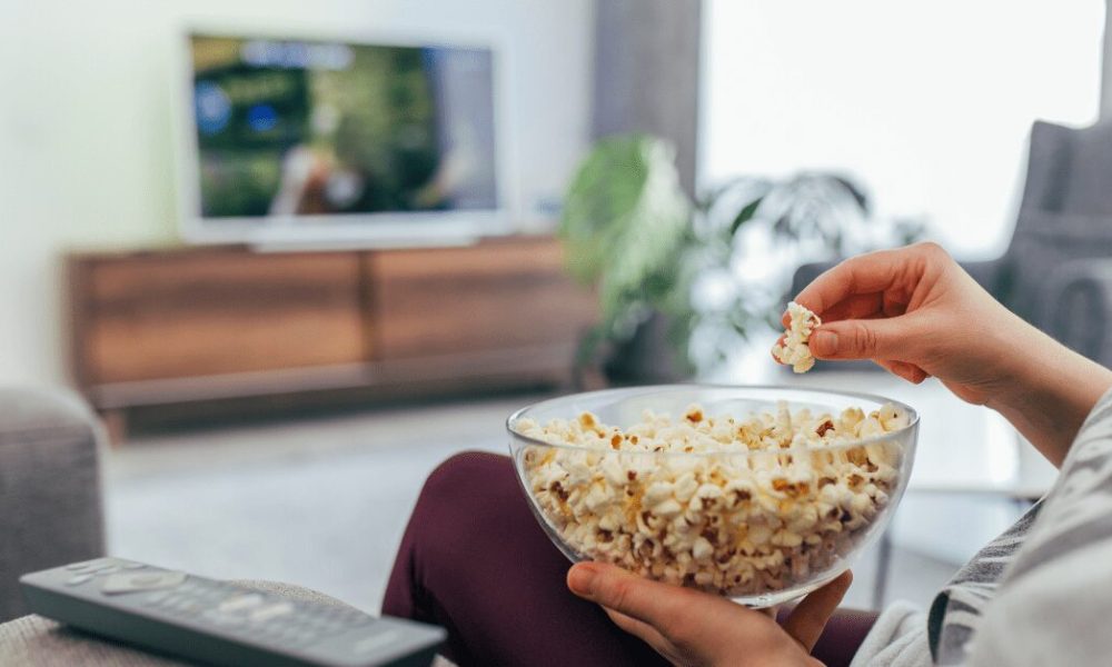 Closeup of hand holding popcorn bowl as a person sits on a couch watching TV.
