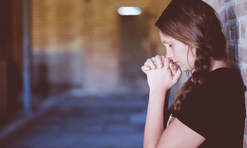 Young woman praying standing up by a wall.